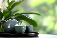 Traditional green ceramic teapot and cups set on dark wooden tray against blurred natural background with white orchid flower and leaves, zen style composition.