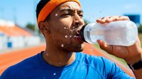 Male athlete of African descent wearing orange headband and blue shirt drinking water from bottle during outdoor training session on sunny day.
