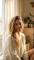 Young blonde woman in cozy cream sweater smiling warmly in sunlit living room with houseplants and natural home decor for lifestyle and wellness content.