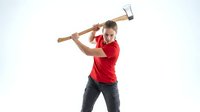 Young Caucasian man in red shirt swinging wooden axe with both hands in chopping motion against white background for lumberjack and woodworking concepts.