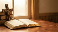 Open Bible and wooden cross on a rustic table by a sunlit window, evoking faith, spirituality, and Christian devotion.