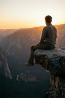 Young man sitting on cliff edge overlooking mountain valley at golden hour sunset, contemplating nature and freedom in peaceful solitude.