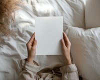 Female hands holding blank white paper sheet on bed with white linen and curly hair visible. Minimalist lifestyle composition in neutral tones.