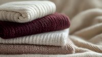 Stack of folded cozy knitted sweaters in neutral beige, burgundy and white colors on soft textile background, warm winter clothing arrangement.