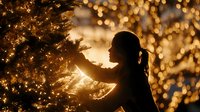 Silhouette of child decorating Christmas tree with golden bokeh lights creating magical holiday atmosphere in background.