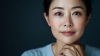 Young Asian woman with gentle smile and thoughtful expression wearing blue sweater against dark background, hand touching chin in contemplative pose.