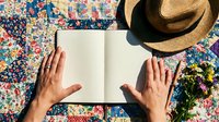 Hands holding open blank book on colorful patchwork quilt with straw hat and flowers, perfect for reading and relaxation concepts.