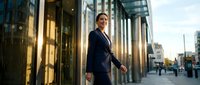 Professional businesswoman in navy suit walking confidently outside modern glass office building during golden hour, representing corporate success and leadership.