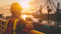Construction worker in safety vest and hard hat overlooking busy port with cranes at golden hour sunset. Industrial maritime logistics and infrastructure development.