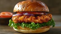 Crispy chicken sandwich with fresh lettuce, red onion, and tomato on glossy brioche bun against dark rustic background, showcasing appetizing fast food presentation.