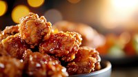 Crispy glazed chicken wings with sweet and spicy Asian sauce, stacked in black bowl against warm bokeh background, close up view of appetizing party food.