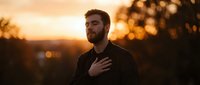 Male adult with reddish-brown beard wearing dark clothing, hand on chest in contemplative pose during golden hour sunset with blurred landscape background.