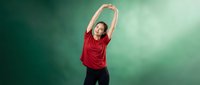 Young athletic woman in red shirt stretching arms overhead during fitness workout routine against green studio background for health and wellness.