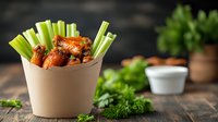Spicy buffalo chicken wings served with fresh celery sticks in brown paper container on rustic wooden table. Creamy dipping sauce and herbs in background.