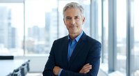 Confident mature Caucasian businessman with gray hair in dark suit standing with crossed arms in modern office with city skyline view.
