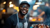 Cheerful African American male chef in glasses and cap smiling while preparing grilled meat at outdoor food station with bokeh lights in background.