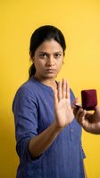 Young Indian woman making stop gesture with hand while holding smartphone on bright yellow background for social media awareness campaigns.