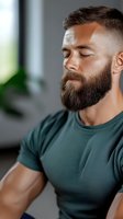 Caucasian male with full dark beard and short hair wearing green t-shirt meditating with eyes closed in peaceful indoor setting. Wellness and mindfulness concept.