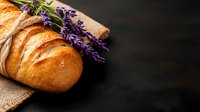 Fresh baked crusty bread loaf with purple lavender flowers on rustic burlap cloth against dark background, food photography with copy space.