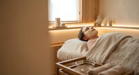 Young woman relaxing peacefully in warm sunlit bedroom with natural lighting streaming through window creating serene morning atmosphere.