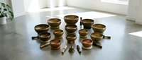 Assorted brass singing bowls arranged in rows on concrete floor with mallets, meditation and sound therapy instruments.