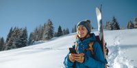 Young woman in blue ski jacket and knit hat holding smartphone on snowy mountain slope with skis and snow-covered pine trees in background.
