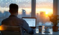 Businessman working on laptop at sunset in modern office with coffee and plant, enjoying peaceful evening atmosphere.