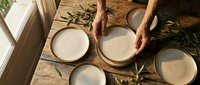 Overhead view of male hands arranging ceramic bowls on wooden table with olive branches and natural lighting for dining or culinary content.