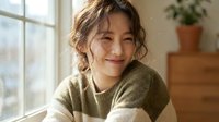 Young Asian woman with wavy brown hair smiling warmly while sitting by window in cozy home interior wearing olive green sweater for lifestyle content.