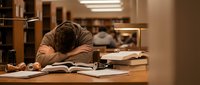 Exhausted student sleeping on desk surrounded by open books and papers in university library during late night study session for exams.