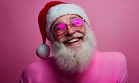 Cheerful senior man with natural white beard wearing Santa hat and pink sweater smiles broadly against pink background, pink-tinted glasses add modern twist.