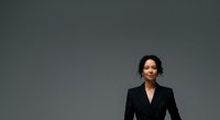 Professional woman with dark wavy hair and olive complexion wearing black blazer against neutral gray background, suitable for corporate, business, and professional headshot applications.