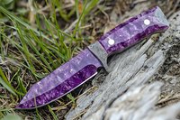 Purple hunting knife with decorative handle lying on weathered wood against green grass background, showcasing detailed craftsmanship and outdoor survival gear styling.