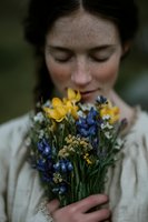 Young woman with freckles smelling wildflower bouquet of yellow and blue flowers in cream sweater for natural beauty and wellness concepts.