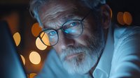 Senior Caucasian man with gray beard and glasses in dramatic blue lighting against blurred bokeh background, contemplative expression and intense gaze.