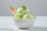 Green pistachio ice cream scoops served in white ceramic bowl with waffle cone on marble surface against light gray background. Food photography.
