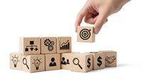 Hand placing wooden cube with target icon among business strategy blocks on white background for corporate planning and goal setting concepts.