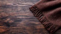 Brown wool scarf with fringe lying on dark wooden surface, creating contrast between soft textile and rustic wood grain texture. Natural lighting highlights materials.