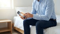 Professional businessman in light blue shirt using smartphone while sitting on bed edge in modern bedroom interior for business communication and mobile technology.