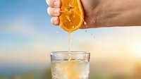 Close-up of hand squeezing fresh orange slice over glass of water with ice against blurred outdoor background, citrus juice dripping into beverage for healthy refreshment.