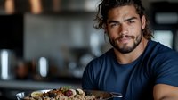 Young Hispanic athletic man with beard smiling while holding healthy meal prep container with grilled chicken, rice and vegetables in modern kitchen setting.