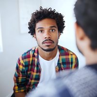 Young African American man with curly hair wearing plaid shirt and white t-shirt listening intently during conversation, professional headshot style portrait.
