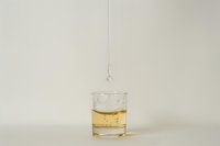 Single drop of water falling into glass of golden liquid on neutral background, creating minimalist composition with subtle ripple effect and light refraction.