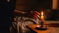 Woman's hand with bracelet reaching over a glowing candle in a holder on a wooden table, warm moody atmosphere in a dark cozy room.