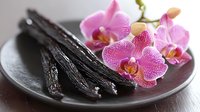 Fresh vanilla pods and blooming pink orchid flowers on dark ceramic plate, aromatic spice and exotic floral composition for culinary and spa concepts.