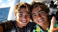 Young Hispanic twin brothers with bright smiles taking selfie outdoors on sunny day. Natural lighting highlights their matching wavy brown hair and joyful expressions.