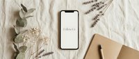 Smartphone with Follow Us text on screen, flat lay with dried flowers, eucalyptus, notebook on linen background, social media concept.