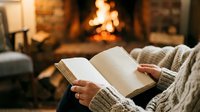 Person reading open book by fireplace in cozy living room with warm blanket, relaxing at home during winter evening.