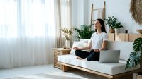 Young Asian woman practicing meditation in modern minimalist living room with plants and natural light streaming through windows.