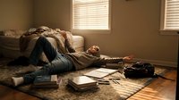 Young woman lying on wooden floor reading book in sunlit bedroom with unmade bed and scattered papers creating cozy morning study atmosphere.